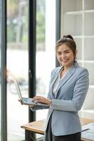 portrait, professionnel et sur de soi asiatique femme d'affaires ou femelle exécutif directeur dans formel costume et utilisation portable. debout, penché sur tableau, en portant portable et en utilisant portable ordinateur. photo