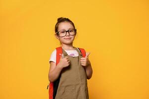 adorable peu enfant fille dans décontractée vêtements et lunettes, porter sa sac à dos, gestes avec les pouces en haut à la recherche à caméra photo