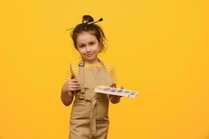 studio portrait peu fille peintre artiste sourit à la recherche à caméra, détient La peinture outils et une palette avec aquarelles photo