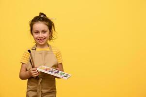 magnifique enfant fille avec peindre brosse et aquarelle palette, inspiré peu peintre, artiste sourit à la recherche à caméra photo