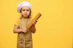 adorable peu enfant fille dans du chef chapeau et tablier, stade avec une roulant broche, isolé plus de Jaune studio Contexte photo