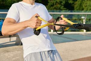 tondu vue de athlète, musclé construire caucasien homme, sportif dans blanc T-shirt Faire force entraînement, exercice avec suspension les bretelles en plein air sur été journée. photo