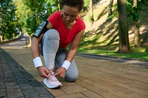 actif âge moyen femme, africain américain athlète attacher les lacets avoir prêt pour faire du jogging le long de le forêt parc sur une chaud ensoleillé été journée. photo