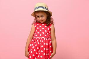 jolie fille dans une rose été robe avec blanc polka points et une paille chapeau, à la recherche timidement à le caméra, posant contre une rose mur Contexte. portrait de une content souriant adorable enfant 3-4 ans vieux. photo