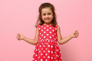 visage portrait de une magnifique enfant fille dans rose polka points robe, mignonne souriant à la recherche à caméra, posant avec les pouces en haut, plus de rose Contexte avec copie espace photo