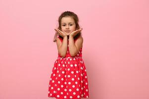 adorable fille dans une rose robe avec polka points pose à le caméra, permanent contre une rose Contexte et en portant sa menton avec sa mains. portrait de une magnifique fille avec positif joyeux émotions photo