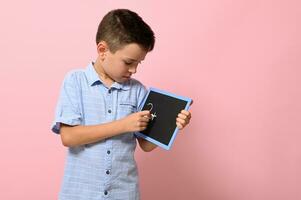 une école garçon dans bleu chemise écrit avec craie sur une tableau noir, isolé plus de rose Contexte avec copie espace. retour à école, concepts photo