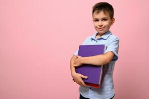 adorable écolier détient livres dans le sien mains une mignonne sourit posant sur caméra, isolé plus de rose Contexte avec espace pour texte. retour à école concepts photo