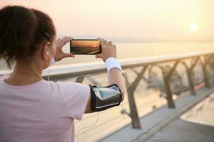 arrière vue de une sportive photographier une lever du soleil pendant sa Extérieur entraînement, permanent sur une tapis roulant sur le Urbain pont, admiratif magnifique la nature Contexte à Aube photo