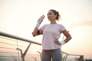 magnifique sportive, femelle athlète les boissons l'eau à hydrater corps après Extérieur cardio entraînement. milieu vieilli athlétique femme repos après le jogging à aube. sport, corps se soucier et actif mode de vie concept photo