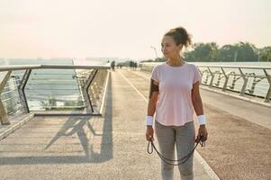 hispanique des sports femme avec une saut corde sur le ville pont. femelle athlète relaxant après intense physique des exercices avec une sauter corde Extérieur. en bonne santé actif mode de vie et cardio faire des exercices concept photo