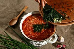 le du chef mains remplir fraîchement bouilli Bortsch avec haché Frais aneth, herbes et Ail. nourriture composition avec une pot de ukrainien traditionnel soupe, en bois cuillère et vert oignons sur une lin nappe de table photo