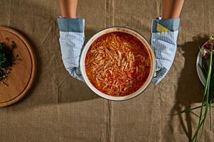 Haut vue de du chef mains dans cuisine gants, en portant pot de fraîchement fabriqué Bortsch soupe et placement il sur table avec lin nappe de table suivant à Coupe planche rempli avec haché aneth et vert oignons photo