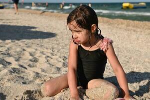une peu brunette fille dans élégant maillot de bain séance sur une sablonneux plage par le mer et en jouant avec le sable. enfant ayant bain de soleil, profiter magnifique ensoleillé journée pendant été vacances photo