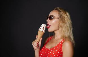 côté portrait de une content blond femme portant une rouge maillot de bain avec blanc polka points et des lunettes de soleil en mangeant une cornet glace, isolé plus de noir Contexte avec copie espace pour publicité photo