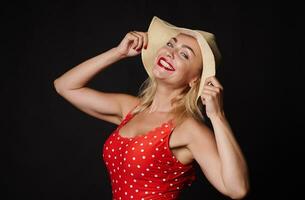 côté portrait de une magnifique joyeux blond femme portant une rouge maillot de bain avec blanc polka points et une été chapeau, souriant avec de bonne humeur blanc et en bonne santé à pleines dents sourire posant contre noir Contexte photo