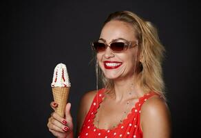 de bonne humeur milieu vieilli blond femme portant protecteur des lunettes de soleil et rouge maillot de bain, en portant une crème glacée cône et sourit à pleines dents sourire à la recherche à caméra, isolé plus de noir Contexte avec copie espace photo