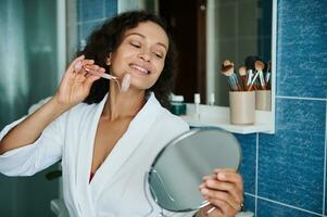 souriant africain américain femme masser sa visage avec une jade rouleau masseur et à la recherche à sa réflexion dans une petit rond miroir photo
