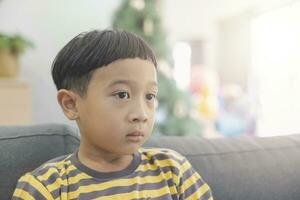 fermer asiatique garçon avec noir cheveux portant une jaune-gris rayé T-shirt à la recherche à le la télé dans le Contexte de Noël des arbres et en bois meubles. enfant fait du marrant visages dans le sien maison Heureusement. photo