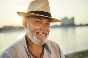 portrait de une souriant Sénior homme portant été chapeau à le au bord de la rivière à le coucher du soleil photo