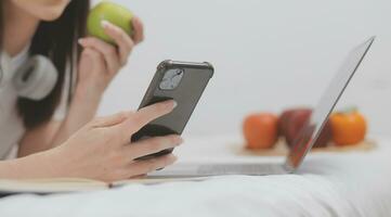 efficace Jeune millénaire fille séance sur une lit dans le matin, les usages portable ordinateur et mange des croissants et les boissons café pour petit déjeuner photo