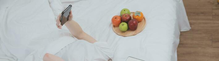 efficace Jeune millénaire fille séance sur une lit dans le matin, les usages portable ordinateur et mange des croissants et les boissons café pour petit déjeuner photo