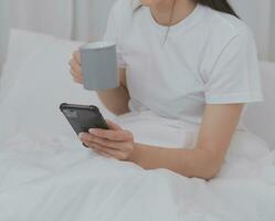 efficace Jeune millénaire fille séance sur une lit dans le matin, les usages portable ordinateur et mange des croissants et les boissons café pour petit déjeuner photo