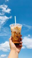 main en portant glacé café dans une Plastique tasse avec une bleu ciel et nuage Contexte. ai généré photo