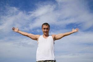 homme dans une blanc T-shirt posé le sien mains large contre le bleu ciel avec des nuages. photo