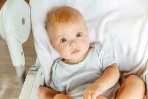 mignonne peu nouveau née fille avec souriant visage à la recherche à caméra sur blanc Contexte. bébé bébé repos en jouant mensonge vers le bas sur alimentation chaise à maison. maternité content enfant concept. photo