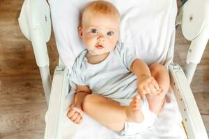 mignonne peu nouveau née fille avec souriant visage à la recherche à caméra sur blanc Contexte. bébé bébé repos en jouant mensonge vers le bas sur alimentation chaise à maison. maternité content enfant concept. photo