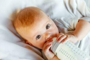 mignonne peu nouveau née fille en buvant Lait de bouteille et à la recherche à caméra sur blanc Contexte. bébé bébé succion en mangeant Lait nutrition mensonge vers le bas sur lit de bébé lit à maison. maternité content enfant concept. photo