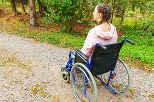 jeune femme handicapée heureuse en fauteuil roulant sur la route dans le parc de l'hôpital en attente de services aux patients. fille paralysée dans une chaise invalide pour personnes handicapées en plein air dans la nature. notion de réhabilitation. photo