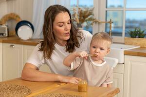 content famille à maison. mère alimentation bébé dans cuisine. peu garçon avec désordonné marrant visage mange en bonne santé aliments. enfant apprend manger par lui-même en portant cuillère. femme maman donnant nourriture à enfant fils. soi alimentation. photo