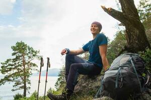 randonnée escalade falaise aventure. randonneur homme à la recherche à magnifique voir. promeneur avec sac à dos est assis sur gros Roche au dessus vert forêt et lac. Jeune content promeneur homme souriant prendre plaisir une randonnée trekking tourisme. photo