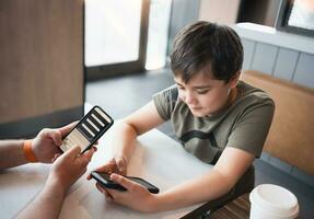 Jeune garçon séance avec parent en utilisant mobile téléphone, mec main en portant intelligent téléphone commande nourriture dans restaurant,enfant en jouant Jeu ou envoyer des SMS messages sur cellule téléphone à amis.technologie et mode de vie concept photo