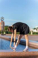 jeune, en forme et sportif fille dans noir vêtements élongation après le faire des exercices près le Publique Fontaine dans le Urbain ville parc. photo