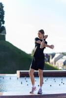 jeune, en forme et sportif fille dans noir vêtements élongation après le faire des exercices près le Publique Fontaine dans le Urbain ville parc. photo