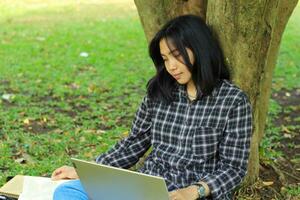 portrait de concentré Jeune asiatique femme surfant l'Internet en utilisant portable à travail à distance, lis livre, et l'écriture dans carnet. attrayant asiatique étudiant étude dans en plein air photo