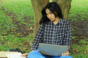 portrait de concentré Jeune asiatique femme surfant l'Internet en utilisant portable à travail à distance, lis livre, et l'écriture dans carnet. attrayant asiatique étudiant étude dans en plein air photo
