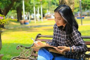 souriant asiatique magnifique Jeune femme prendre plaisir l'écriture à faire liste et idée dans carnet dans en plein air ville parc photo