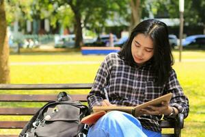 magnifique asiatique Jeune femme Université étudiant concentré l'écriture sur carnet et en train de lire livre dans en plein air ville parc photo