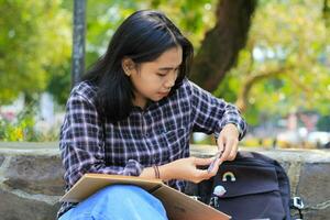 magnifique asiatique Jeune femme Université étudiant concentré l'écriture sur carnet et en train de lire livre dans en plein air ville parc photo