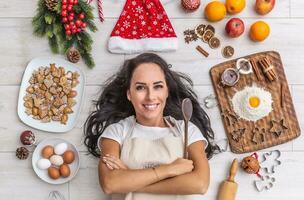 magnifique foncé aux cheveux cuisinier pose et largement souriant sur le sol, en portant le en bois cuillère et étant entouré par pains d'épice, œufs, farine, Noël chapeau, séché des oranges et cuisson formes photo