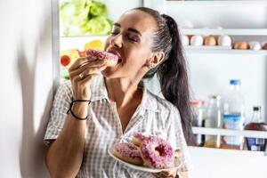 faim brunette dans pyjamas jouit sucré beignets en retard à nuit par le ouvert réfrigérateur photo