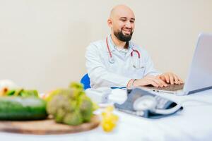 souriant nutritionniste avec portable à bureau. nutritionniste médecin en utilisant portable à lieu de travail. barbu nutritionniste médecin travail sur portable à bureau. photo