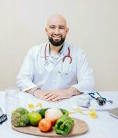 souriant nutritionniste médecin à bureau avec portable et des légumes. barbu nutritionniste médecin à le sien lieu de travail, portrait de souriant nutritionniste à sa bureau photo