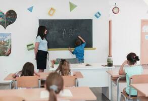 élémentaire école. le femelle prof portion le enfant étudiant tandis que l'écriture le répondre sur le tableau noir. photo