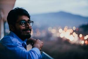 un Indien homme avec des lunettes et une bleu chemise regards autour le ville à nuit. dans le Contexte de le nuit rue de le ville photo