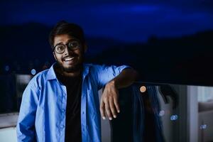 un Indien homme avec des lunettes et une bleu chemise regards autour le ville à nuit. dans le Contexte de le nuit rue de le ville photo